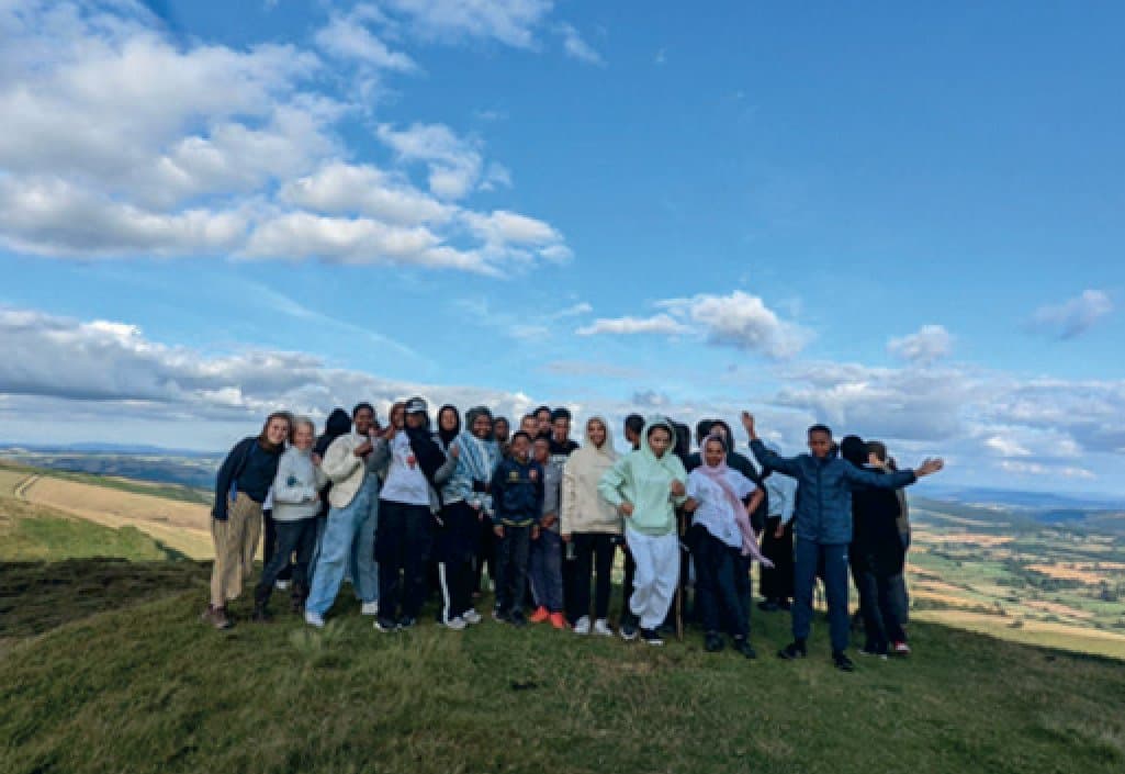 A large group of young people from Minority Matters smiling and waving on a grassy hilltop on a trip
